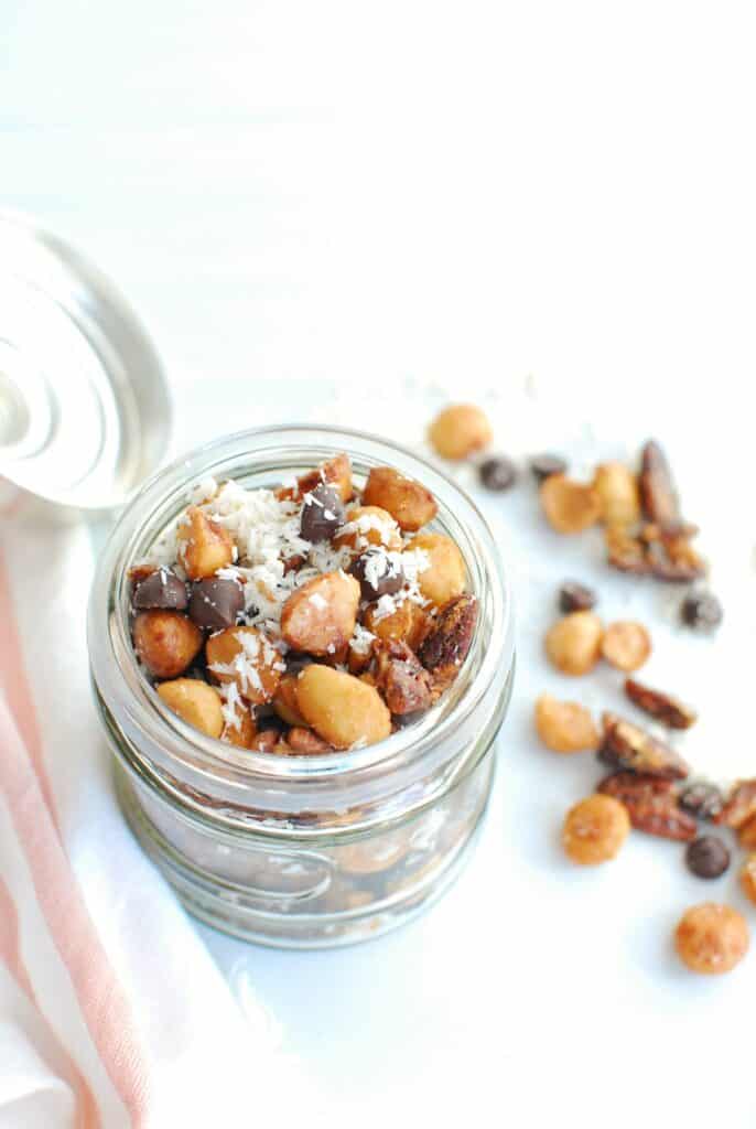 a small glass jar filled with keto trail mix, next to some chocolate chips and nuts a small glass jar filled with keto trail mix, next to some chocolate chips and nuts