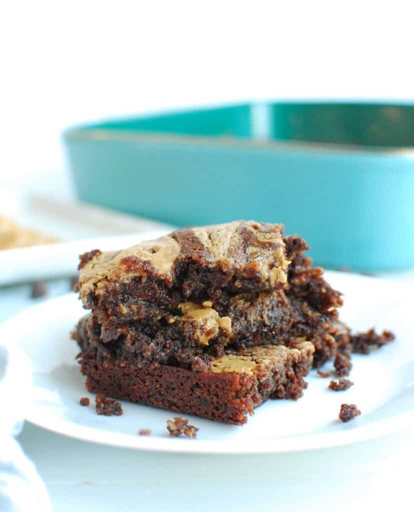 a stack of three large keto peanut butter brownies on a white plate next to the baking dish of brownies a stack of three large keto peanut butter brownies on a white plate next to the baking dish of brownies